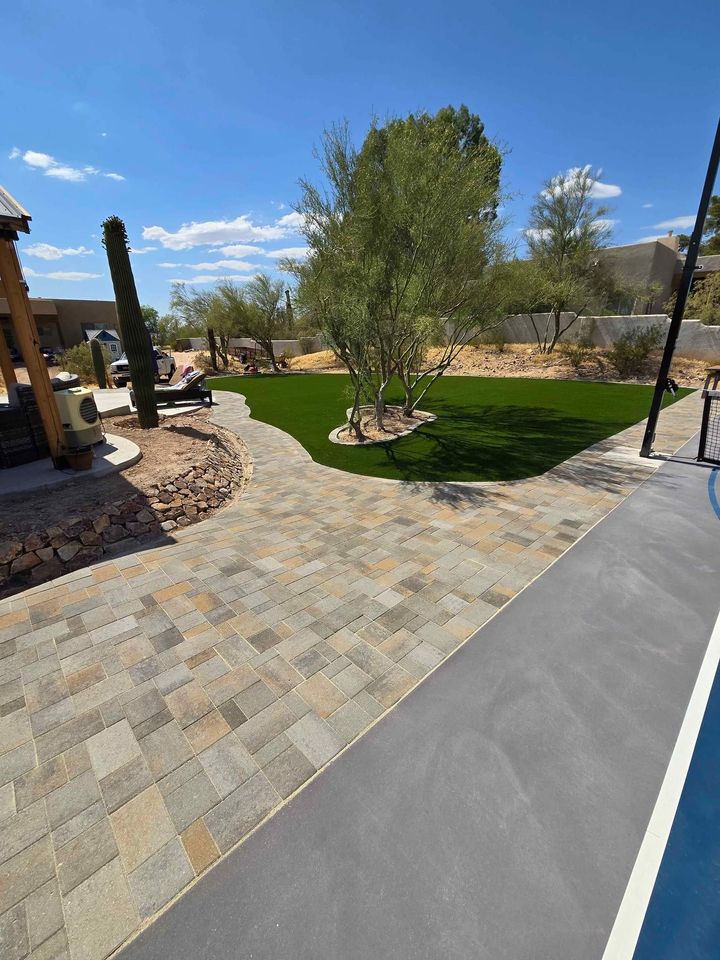 A backyard with paved stone walkway, Hawaii Artificial Turf green lawn, desert plants, and trees, set against a dry landscape with rocks and clear blue sky. Part of a pool and patio area are visible in the foreground.