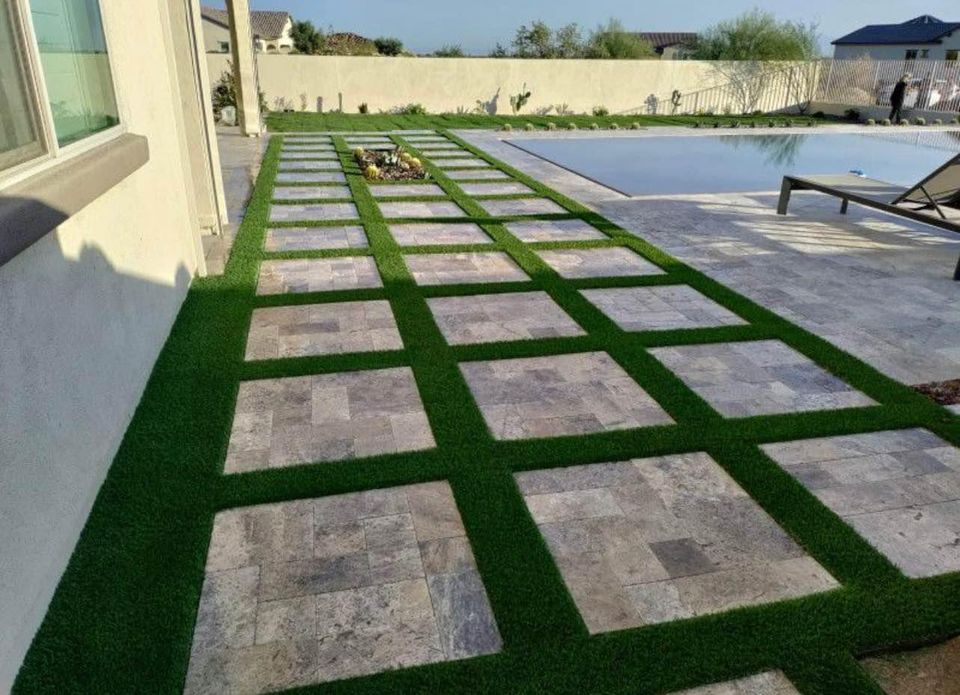 A modern backyard features a pool and a patio with square stone tiles arranged in a grid, separated by strips of lush Hawaii Artificial Turf. Two lounge chairs sit near the pool.