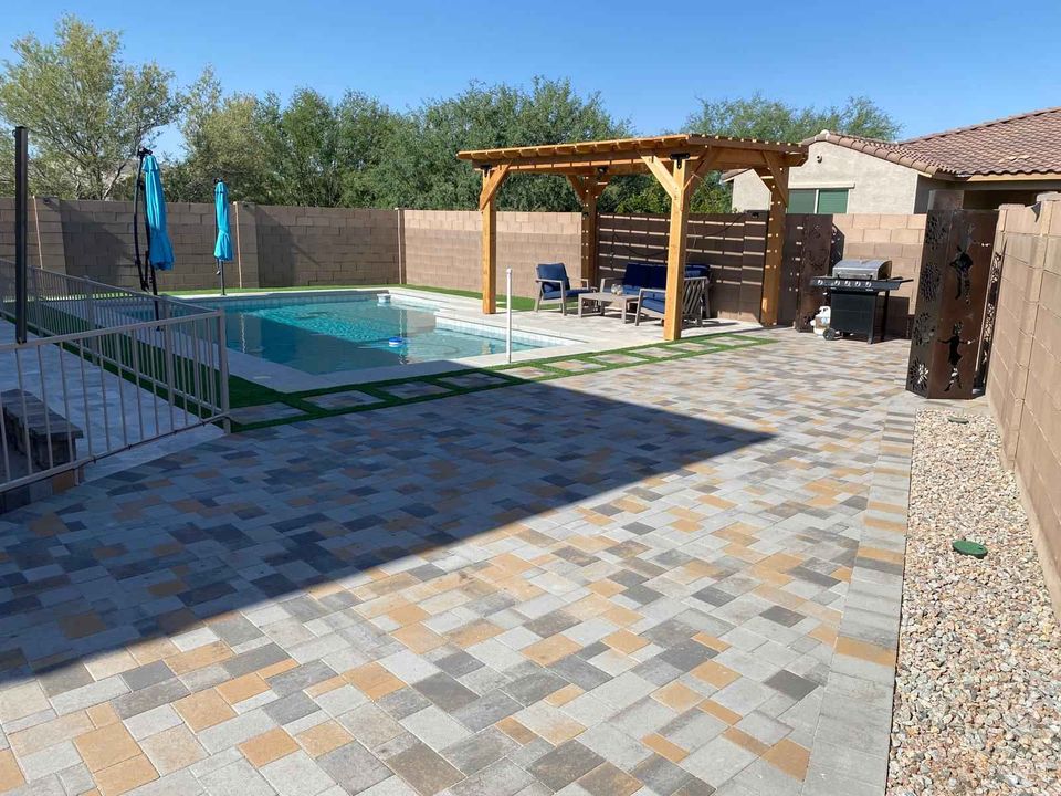 A backyard with a tiled patio, a pergola with outdoor seating, Hawaii Artificial Turf alongside a swimming pool with a safety fence, two blue umbrellas, a grill, and desert landscaping surrounded by a tan brick wall.