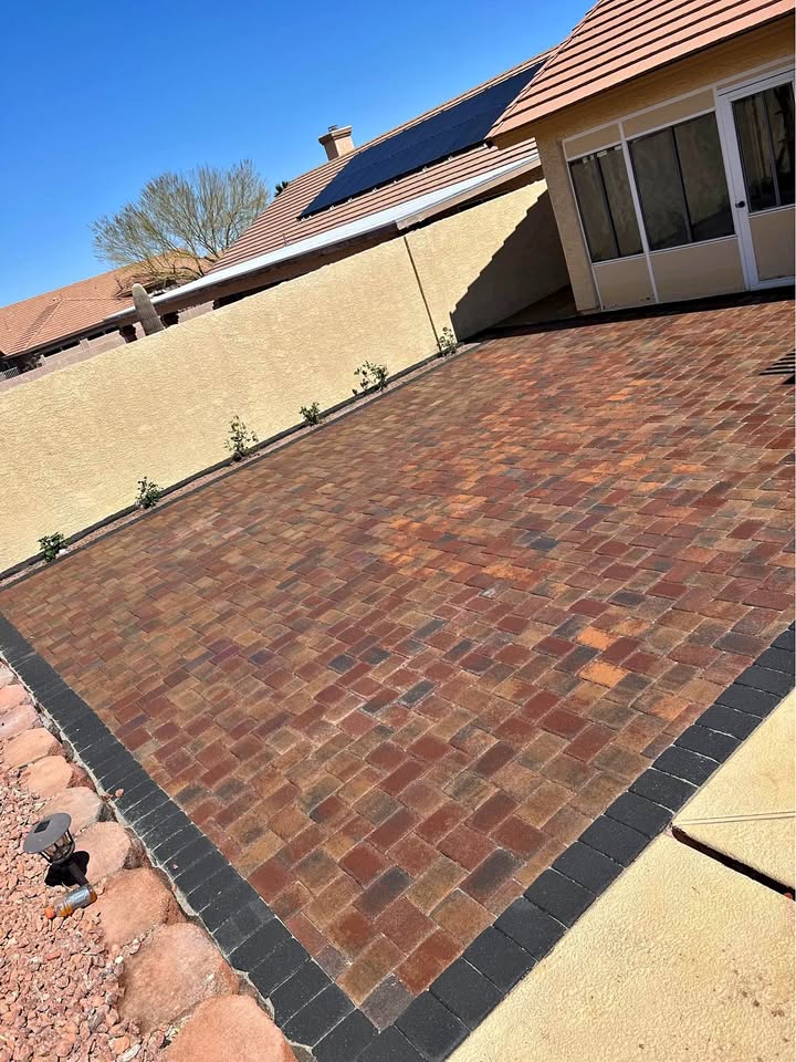 A backyard with a large patio made of reddish-brown and dark pavers, bordered by tan walls. There are small plants along the wall, Hawaii Artificial Turf adding greenery, and a desert landscape with rocks to the side. A house with solar panels is visible.
