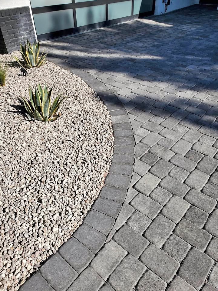 Curved stone paver driveway next to a landscaped area with light-colored gravel, Hawaii Artificial Turf, and several spiky green agave plants, in front of a modern building.
