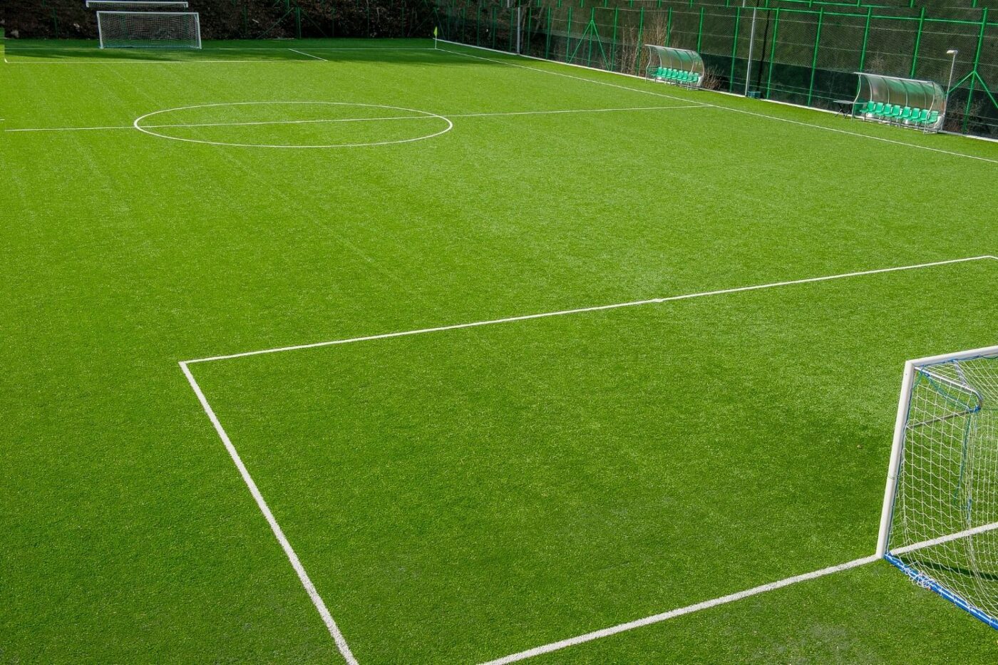 A well-maintained, empty Hawaii Artificial Turf soccer field with crisp white boundary lines, two goals, and green benches along the sideline. The field is surrounded by fencing and trees in the background.