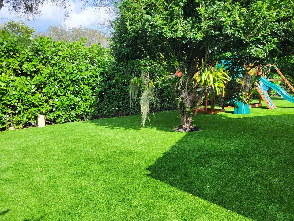 A lush green backyard with neatly trimmed grass, a large tree with hanging plants, dense hedges, and a colorful playground set with slides in the background. Sunlight casts shadows on the lawn.