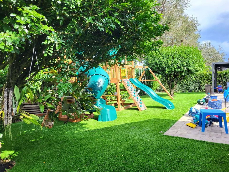 A backyard with lush green grass, a large wooden playset featuring blue slides and a climbing wall, surrounded by trees and plants. There are plastic chairs, a table, and toys on a paved patio area.