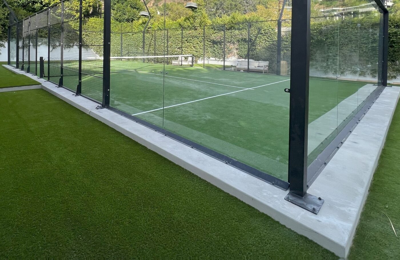 A padel court enclosed by glass walls and surrounded by Sports Field Artificial Turf Oahu HI, with benches along the far side and greenery in the background.
