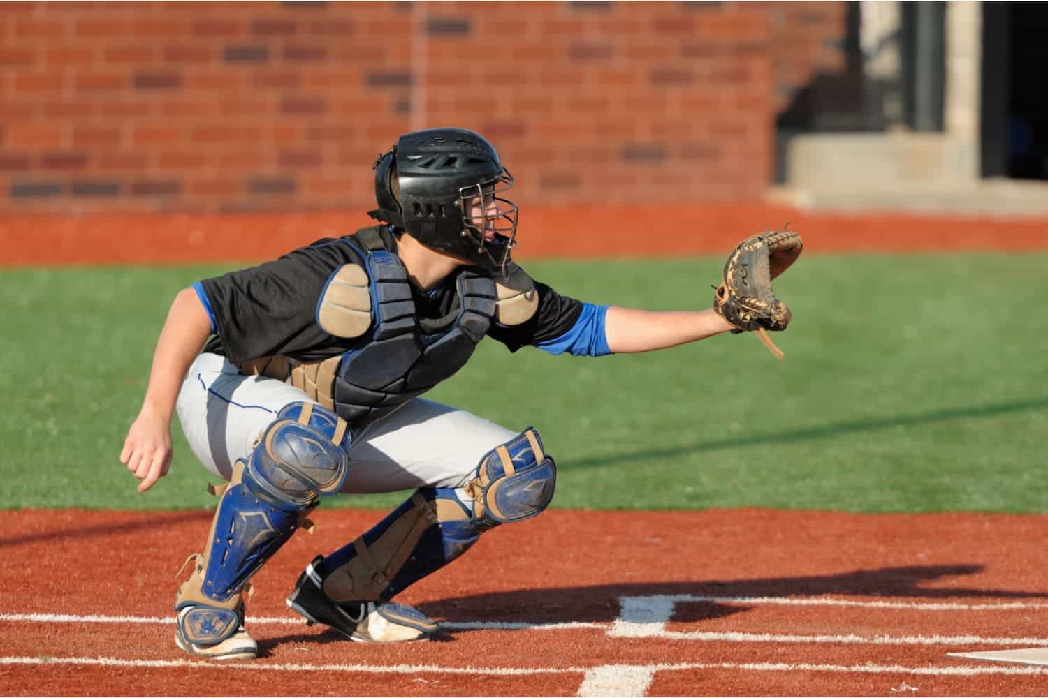 A baseball catcher in full gear squats behind home plate on a sports field with artificial turf in Oahu, HI, wearing a helmet and chest protector, holding a glove out to catch a pitch against a backdrop of green grass and a brick wall.