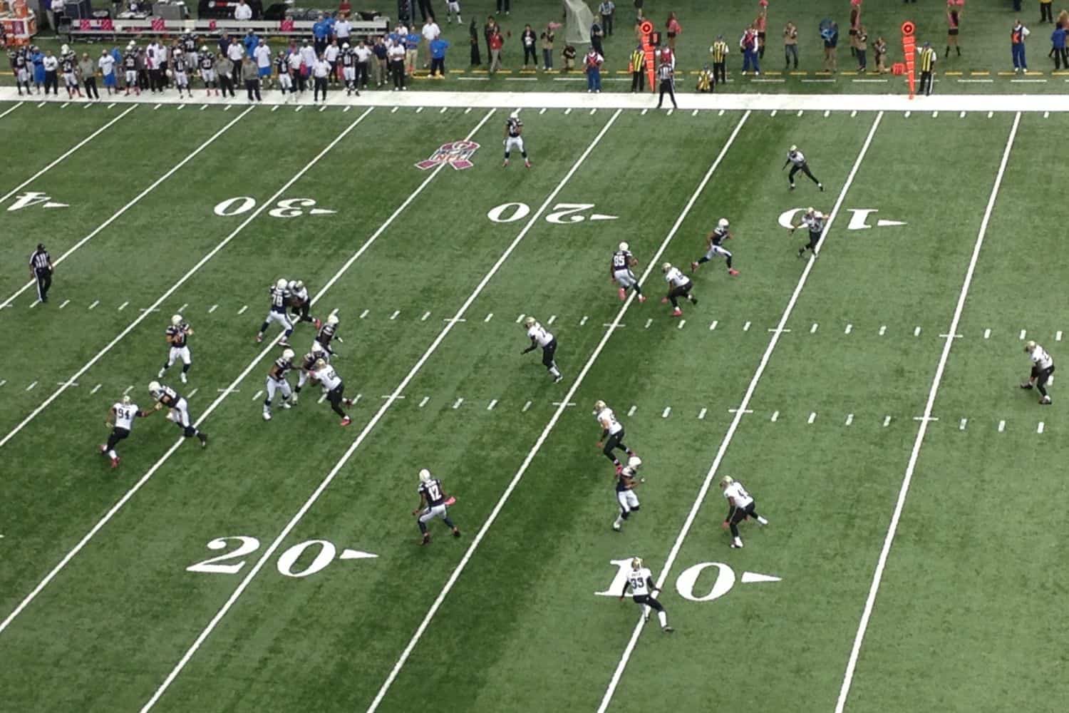 A football game in progress shown from above on a sports field with artificial turf in Oahu, HI. One team is in black and the other in white, lined up near the 20-yard line as spectators watch from the sidelines.