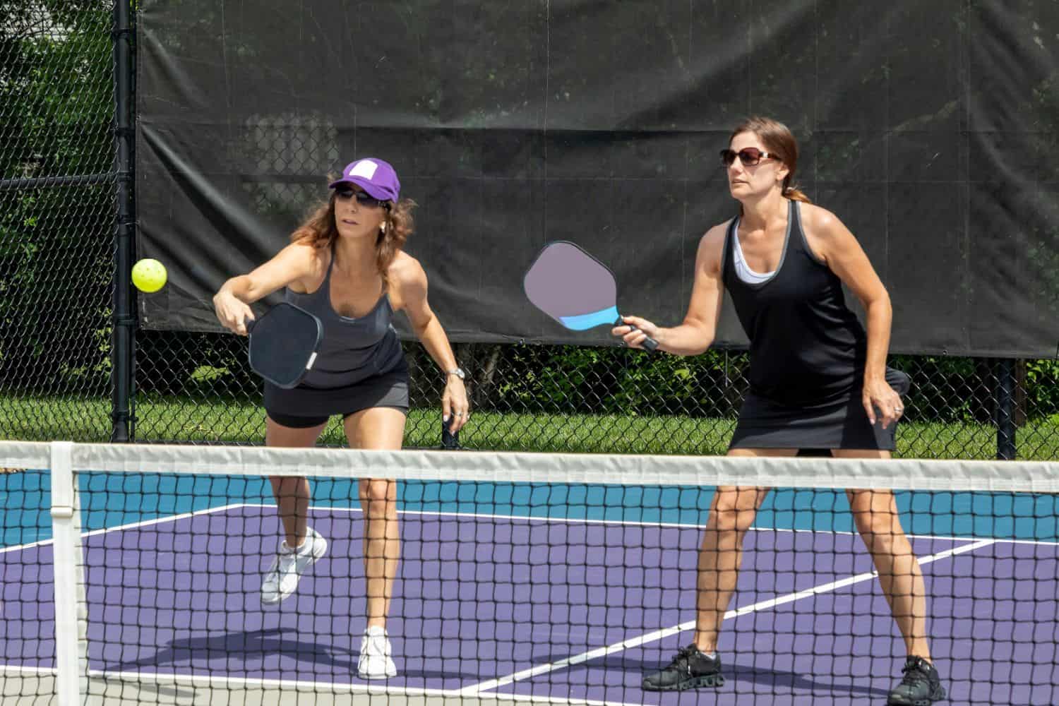 Two women play pickleball on an outdoor court with Sports Field Artificial Turf in Oahu, HI, both holding paddles and focusing intently on a ball in mid-air near the net. A dark fence and greenery complete the scene.