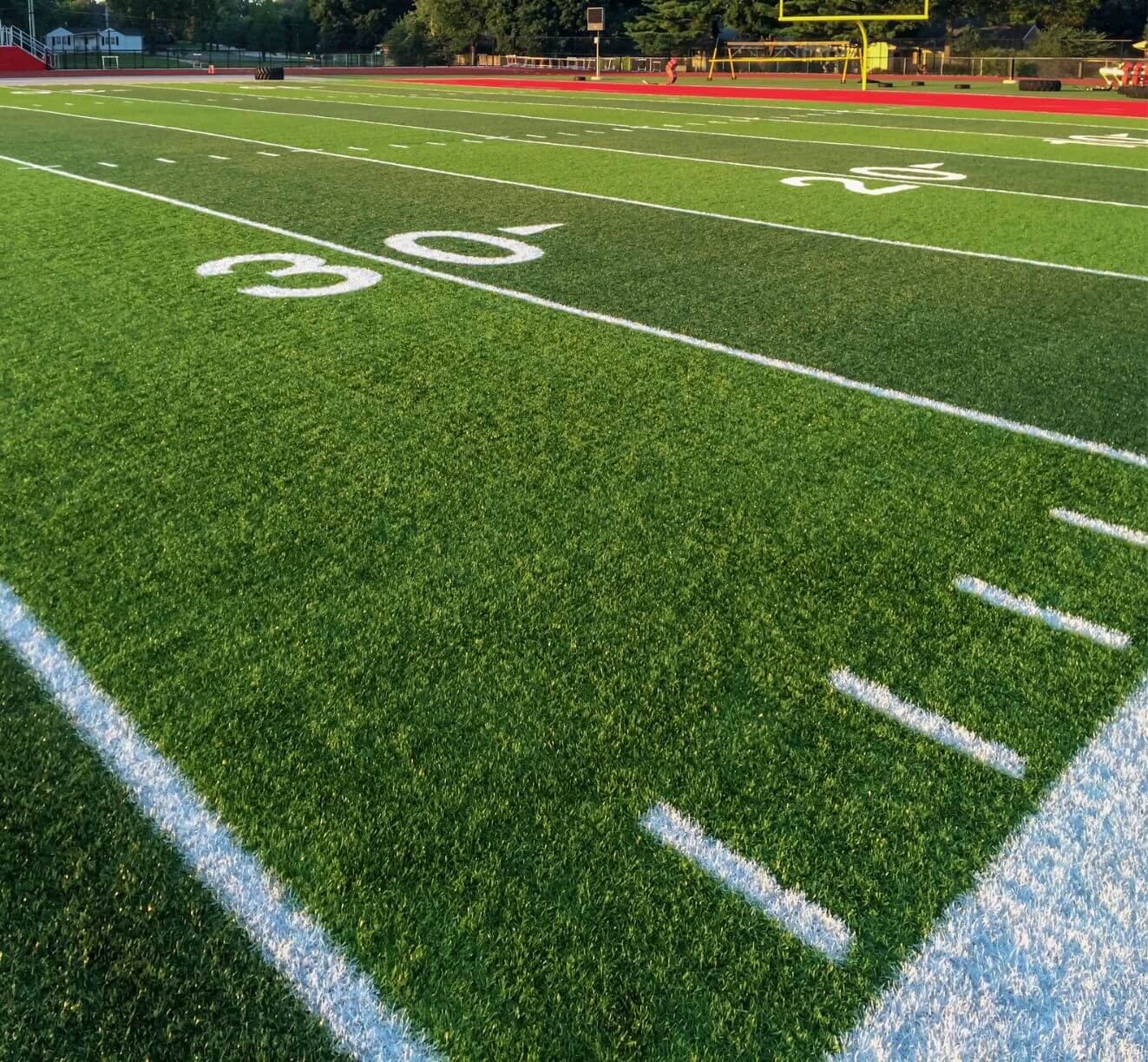 A close-up view of a sports field with crisp white yard lines, numbers, and green artificial turf under sunlight. A yellow goalpost and trees frame this vibrant Oahu, HI football field.
