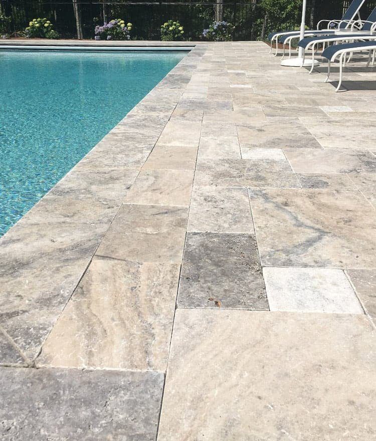 Light-colored stone tiles from a quality paver installation line the edge of an outdoor swimming pool, with several lounge chairs and greenery visible in the background.