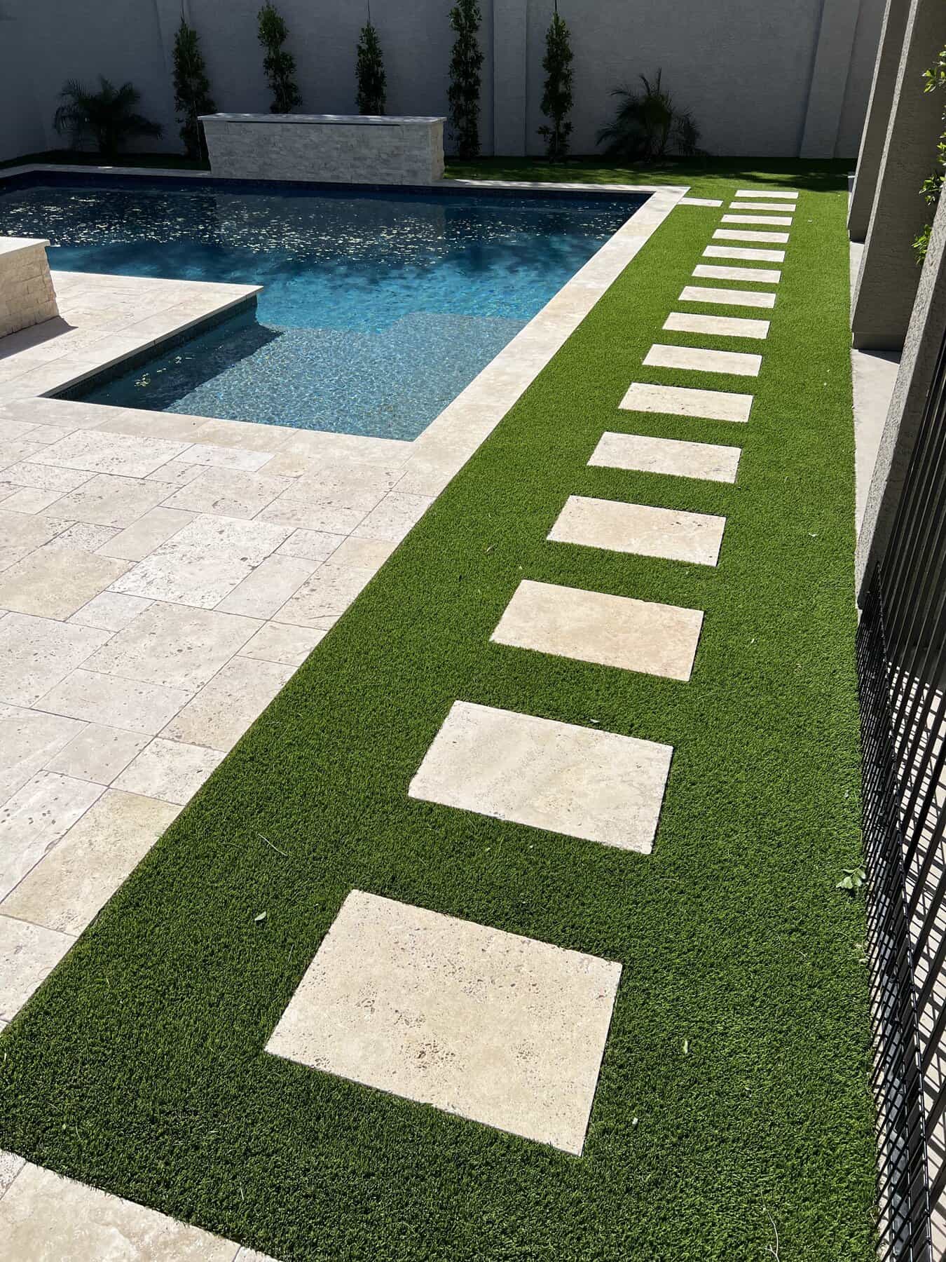 A modern backyard with a swimming pool, beige stone tiles, and lush Hawaii Artificial Turf. Square stone pavers create a path alongside the pool, and there are plants along the far wall.