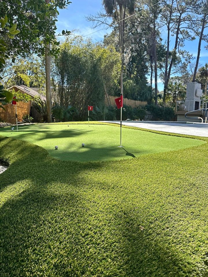 A backyard putting green with Hawaii Artificial Turf features three red flags marking holes. Two golf balls rest nearby, while trees and a paved patio area can be seen in the background beneath a clear sky.