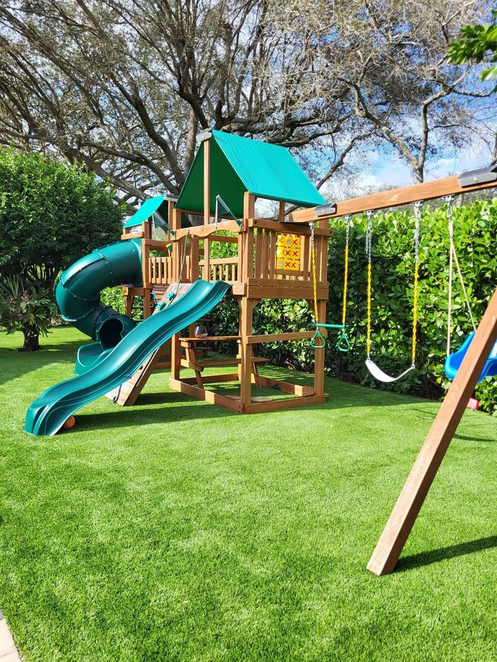 A wooden backyard playset with green slides, swings, a climbing wall, and a covered platform stands on bright Hawaii Artificial Turf, surrounded by bushes and trees under a blue sky.