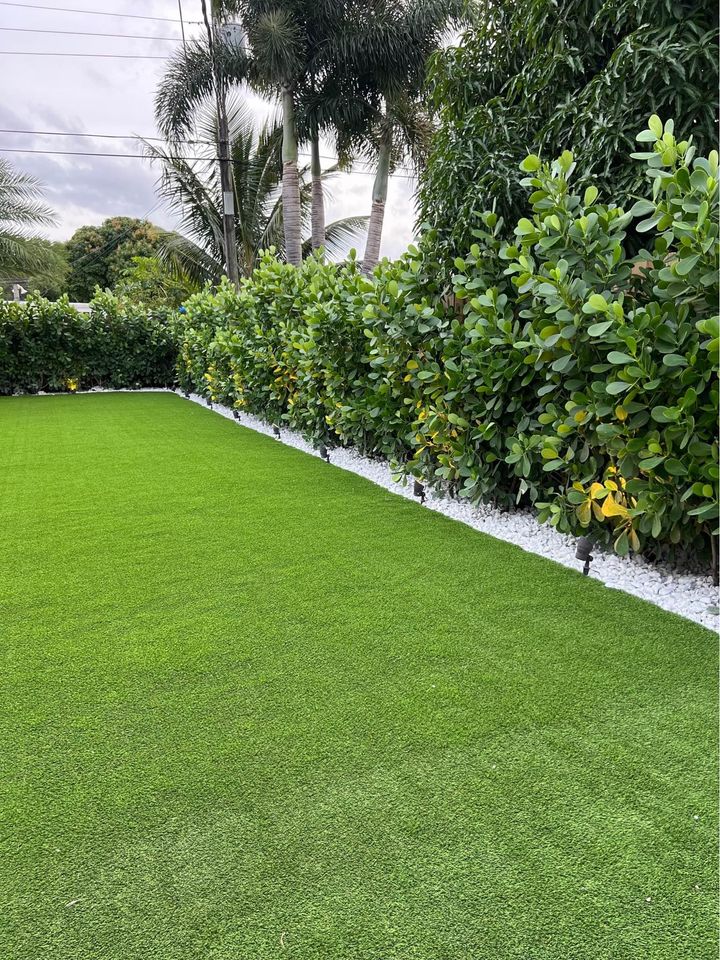 A neatly maintained backyard with bright green Hawaii Artificial Turf, a row of leafy shrubs along the edge, white decorative stones at the base, and tall palm trees in the background under a cloudy sky.