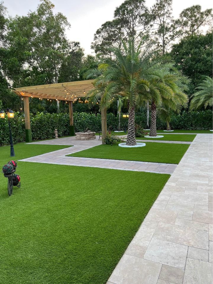 A well-kept garden with lush Hawaii Artificial Turf, palm trees, a paved stone walkway, a wooden pergola with string lights, and a decorative bicycle planter in the corner, surrounded by tall trees and shrubs.