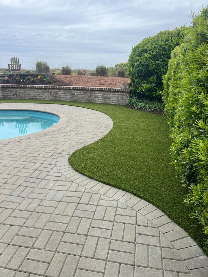A backyard with a curved stone patio surrounding a round pool, bordered by neatly trimmed bushes, lush Hawaii Artificial Turf, and flower beds against a brick wall under a cloudy sky.