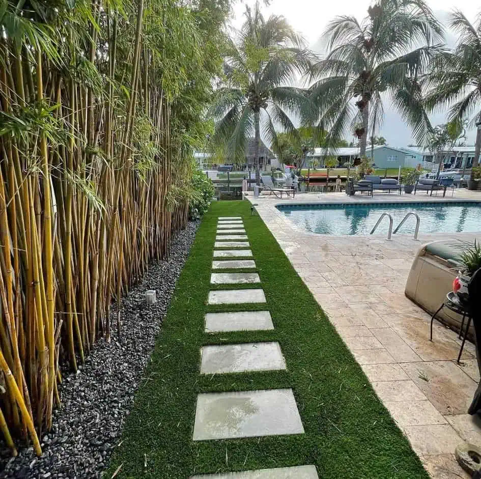 A stone walkway bordered by bamboo, Hawaii Artificial Turf, and black gravel leads to a pool surrounded by palm trees, with lounge chairs and a canal in the background under a partly cloudy sky.