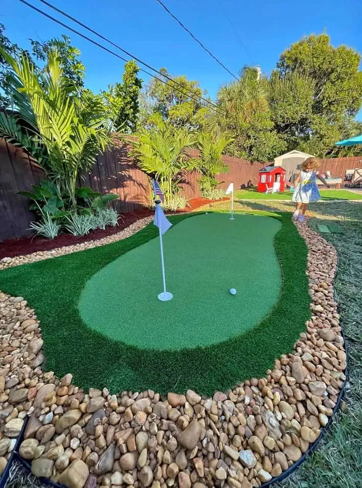 A backyard putting green surrounded by rocks and tropical plants, with two golf flags. A child stands near a small red playhouse in the background on a sunny day.