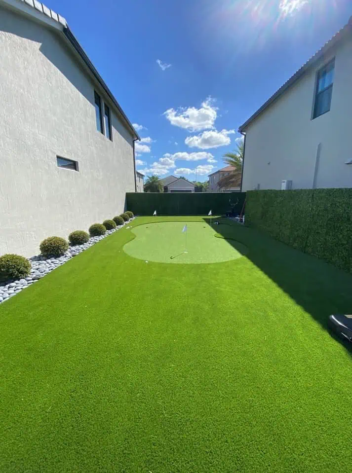 A neatly manicured backyard putting green by Hawaii Artificial Turf lies between two houses, surrounded by artificial grass, small bushes, and decorative rocks under a sunny, blue sky with scattered clouds.