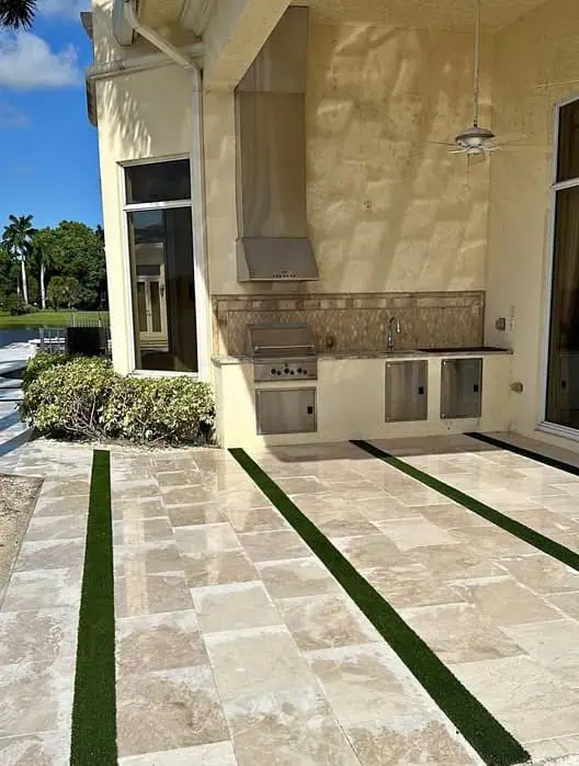 Outdoor patio with light-colored tiles, narrow strips of artificial grass between tiles, a built-in stainless steel grill, sink, and cabinets along the wall, and bushes on the side under a sunny sky.